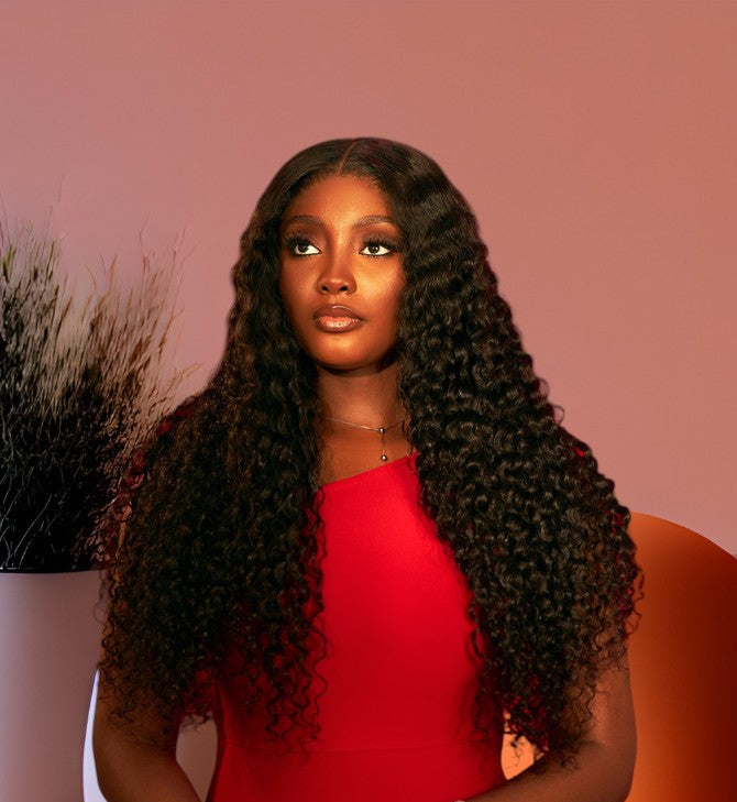 A woman with long, dark, curly hair wearing a bright red one-shoulder dress, standing against a pink studio backdrop with decorative plants and a round orange object in the background.